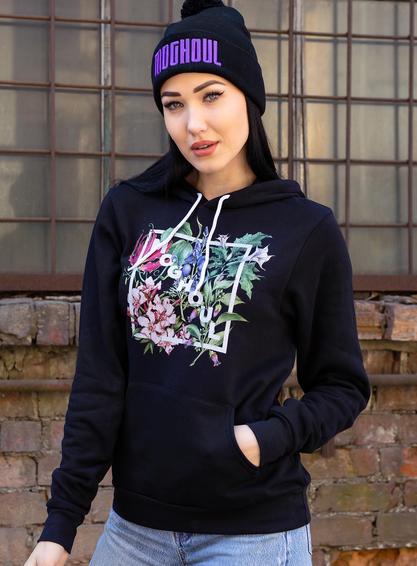 Female modeling a Black unisex hoodie featuring white Moghoul logo surrounded by poisonous flowers such as oleander, fire lily, belladonna and toloache.
