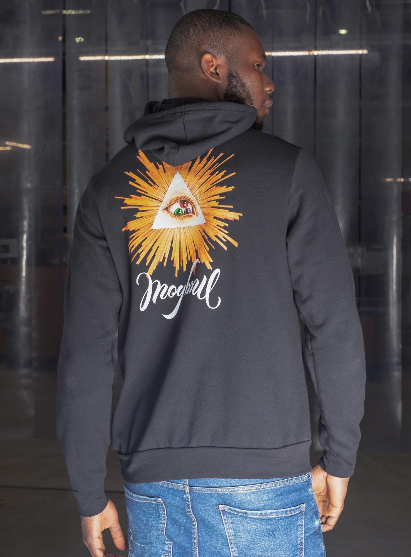 man modeling a black unisex hoodie featuring a back print of a reinterpretation of the Horus pyramid and Moghoul ambigram logo by G.M. Meave.
