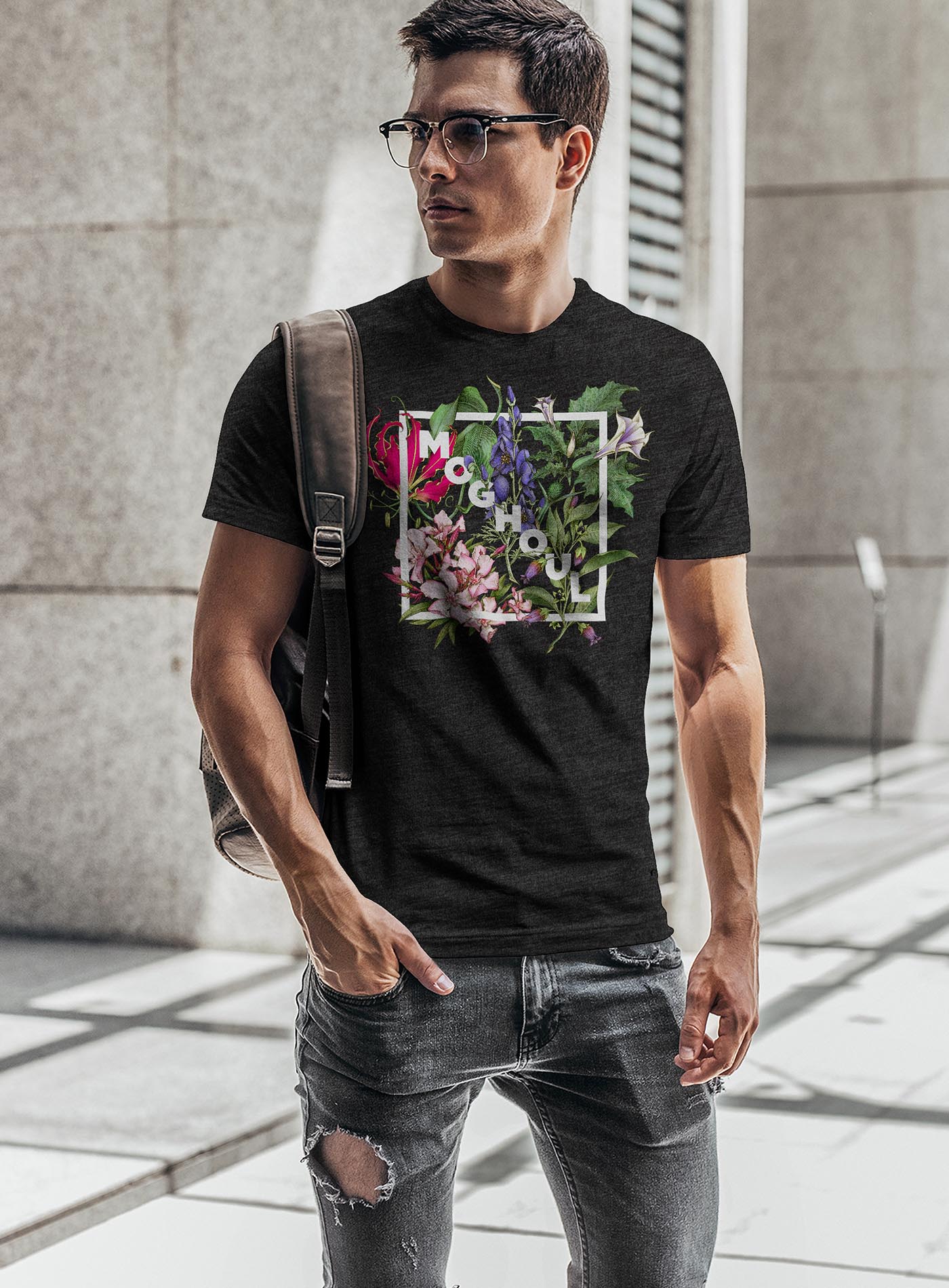 man modeling a Heather black unisex t-shirt featuring white Moghoul logo surrounded by poisonous flowers such as oleander, fire lily, belladonna and toloache.