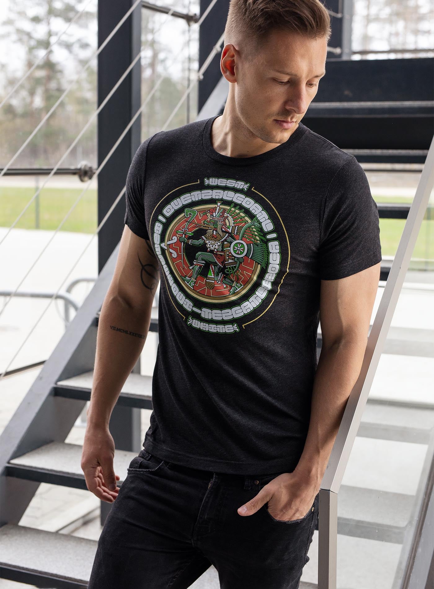 man modeling a Heather black unisex t-shirt featuring a front print of the toltec-aztec god Tezcatlipoca also known as Quetzalcoatl. Reinterpretation by Mexican illustrator G.M. Meave.
