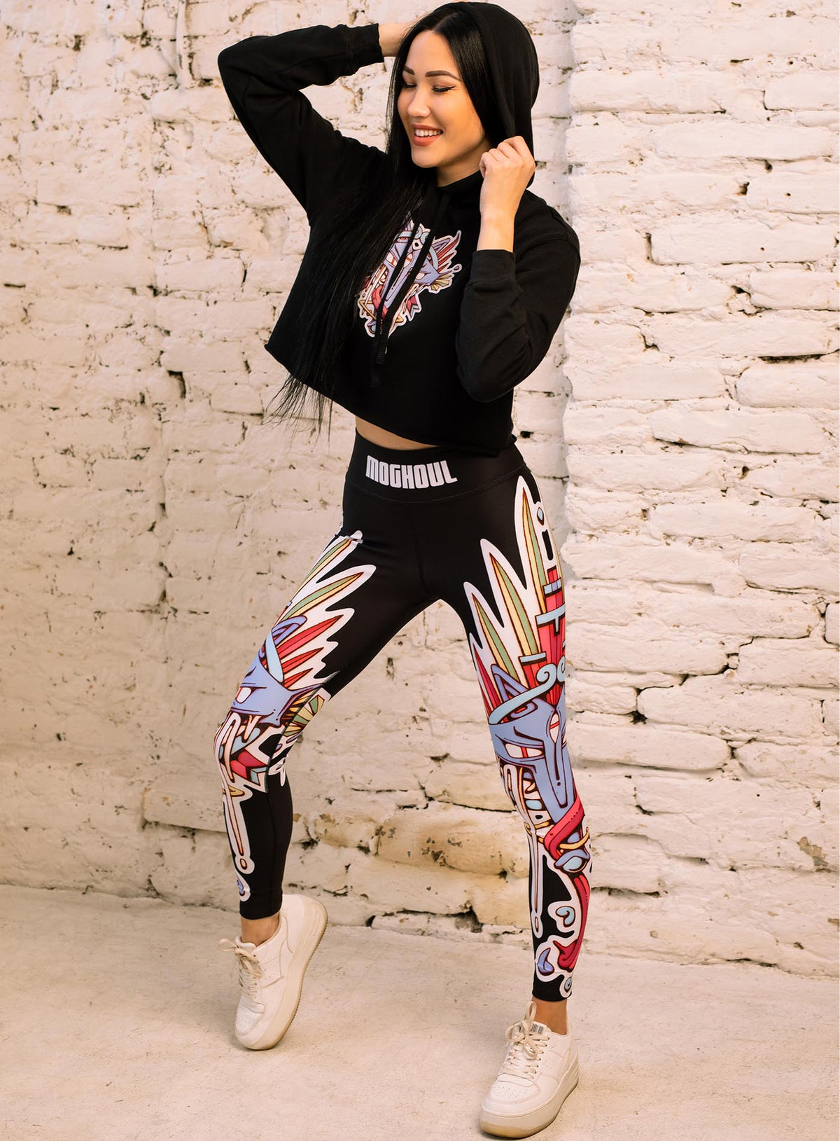 Woman modeling a Black woman&#39;s cropped hoodie and All over dye sublimation leggings featuring a print of the Toltec and Aztec coyote deity illustrated by G.M. Meave.