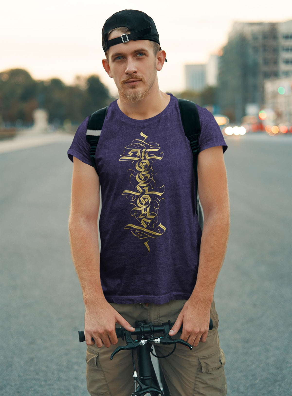 Man modeling a Heather blue unisex t-shirt featuring a front print of a white Moghoul straight-up-gothic logo by Mexican typographer G.M. Meave.