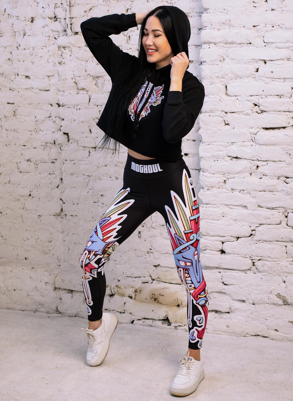 Woman modeling a Black woman&#39;s cropped hoodie and All over dye sublimation leggings featuring a print of the Toltec and Aztec coyote deity illustrated by G.M. Meave.