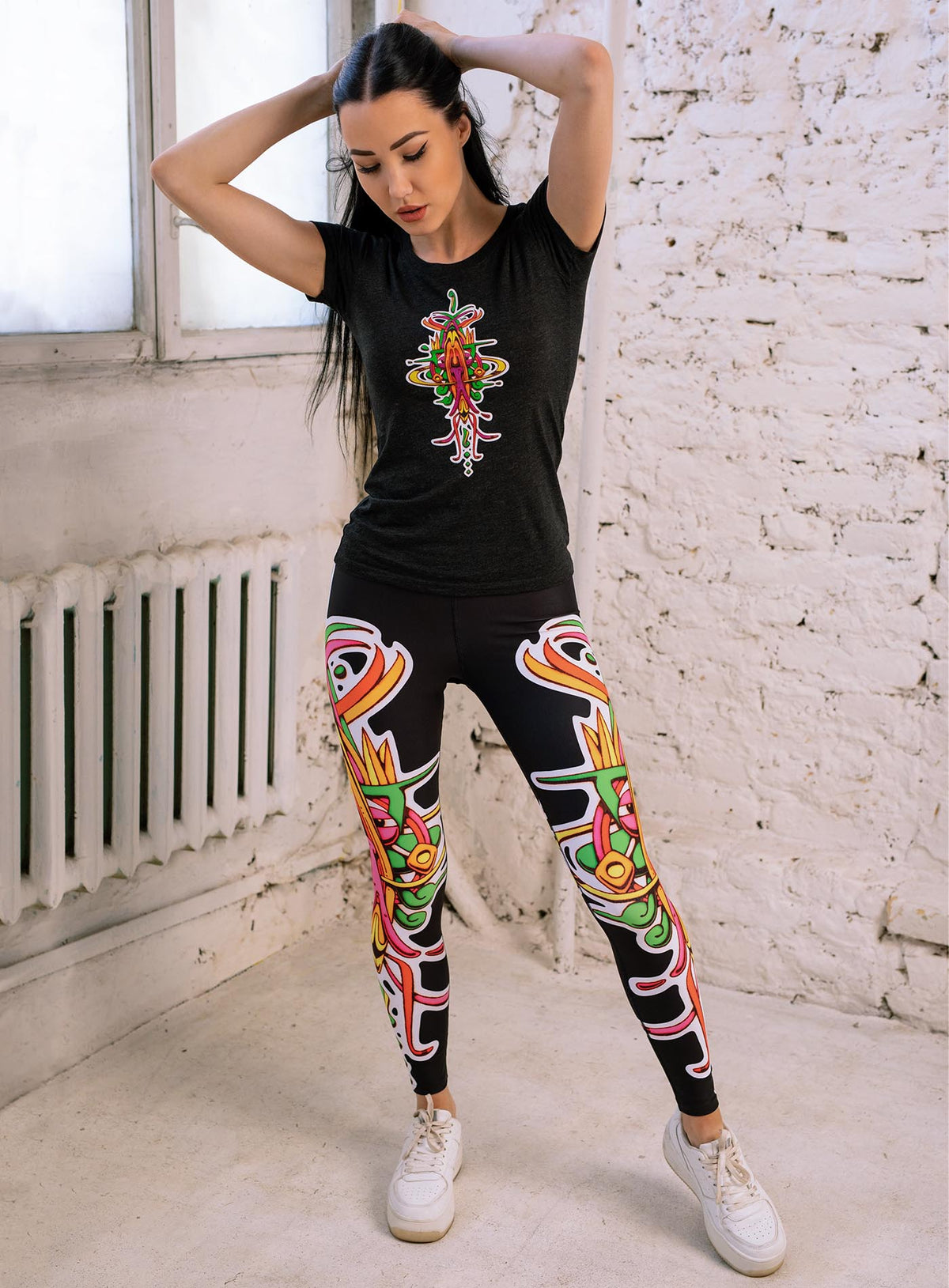 Woman modeling a heather black woman&#39;s t-shirt and all over dye sublimation leggings featuring a front print of the Toltec-Aztec hummingbird deity illustrated by G.M. Meave.