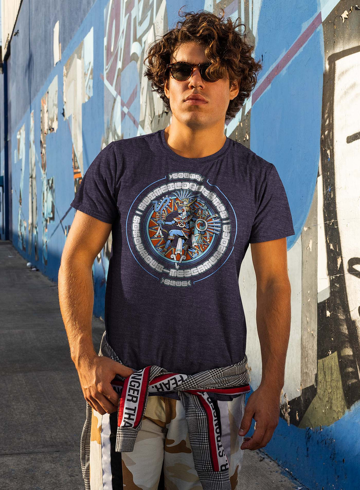 man modeling a Heather blue unisex t-shirt featuring a front print of the toltec-aztec god Tezcatlipoca also known as Huitzilopochtli. Reinterpretation by Mexican illustrator G.M. Meave.