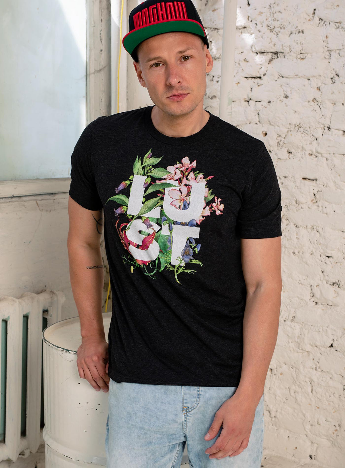 Man modeling a Heather black unisex t-shirt featuring the word LUST surrounded by poisonous flowers such as oleander, fire lily, belladonna and toloache.