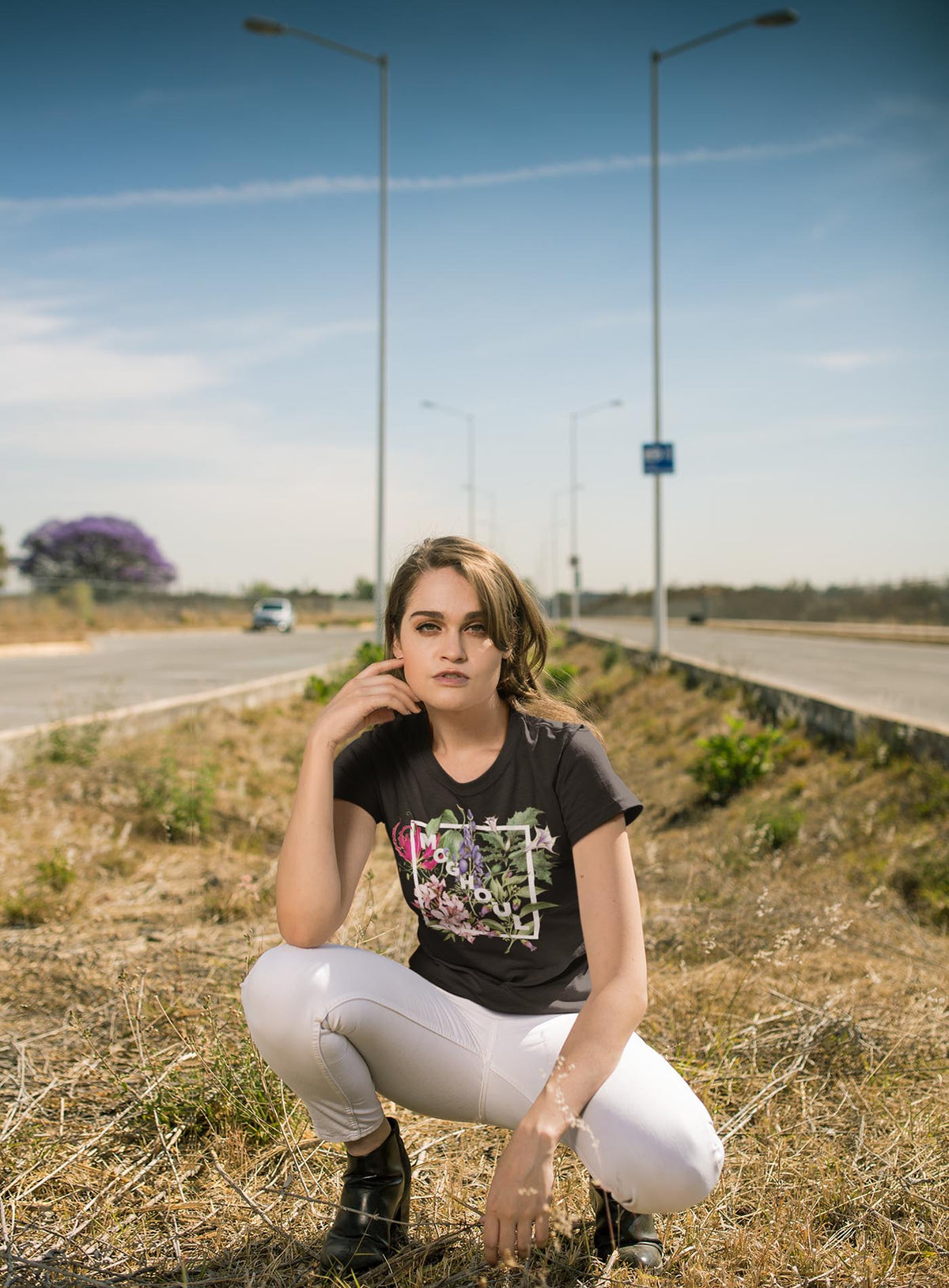 Modeling a Heather black unisex t-shirt featuring white Moghoul logo surrounded by poisonous flowers such as oleander, fire lily, belladonna and toloache.