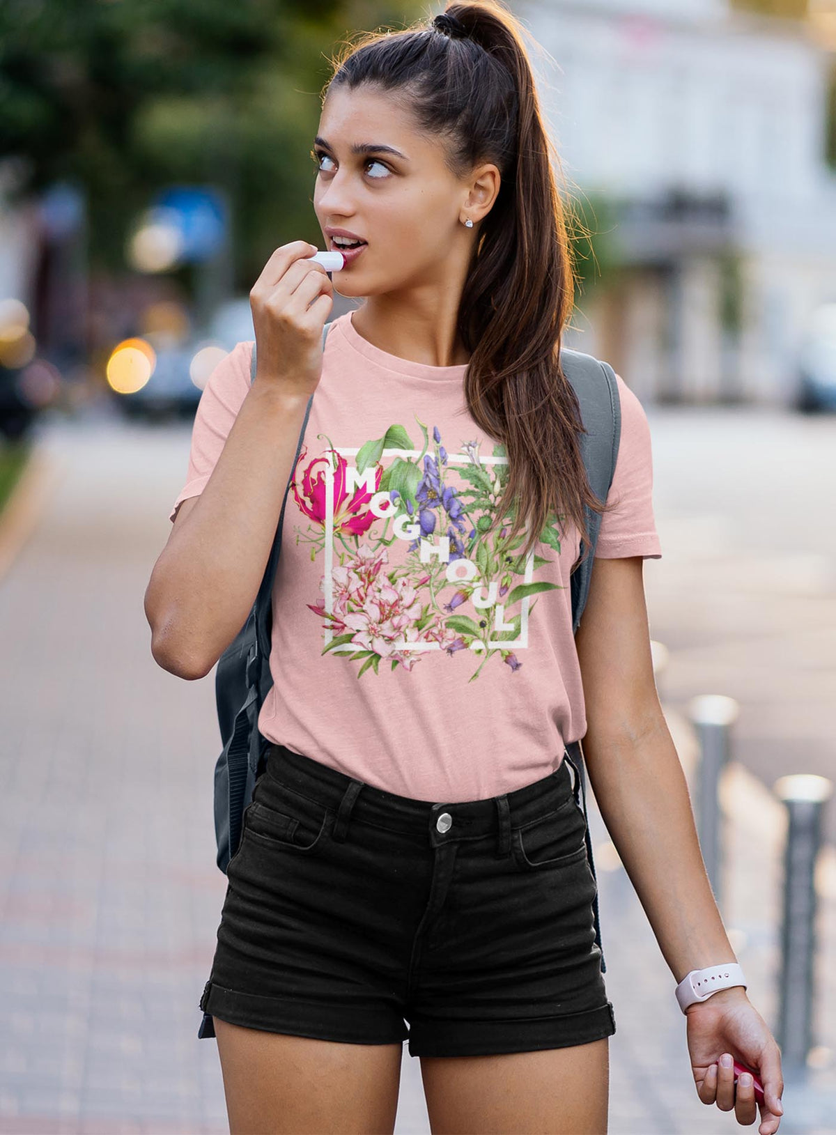 Woman modeling a Pink unisex t-shirt featuring white Moghoul logo surrounded by poisonous flowers such as oleander, fire lily, belladonna and toloache.