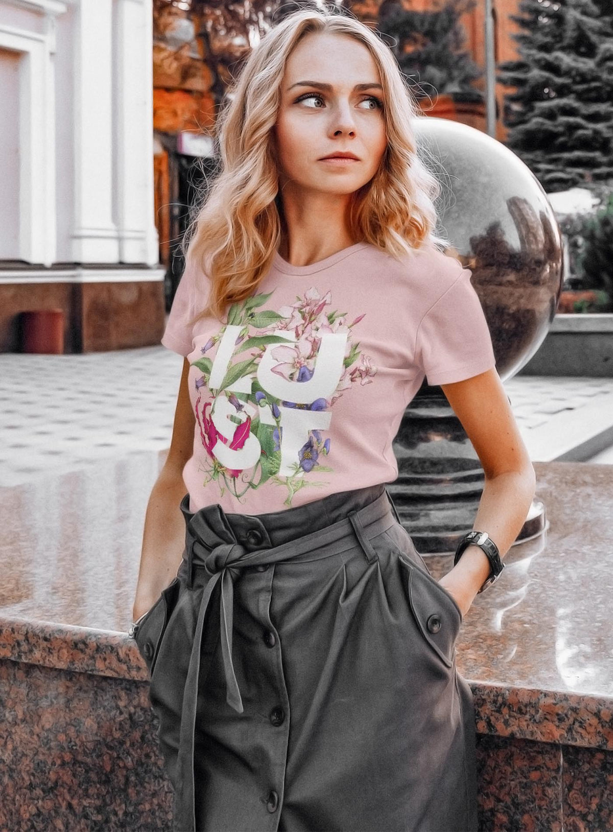 Woman modeling a Detail of Heather pink unisex t-shirt featuring the word LUST surrounded by poisonous flowers such as oleander, fire lily, belladonna and toloache.